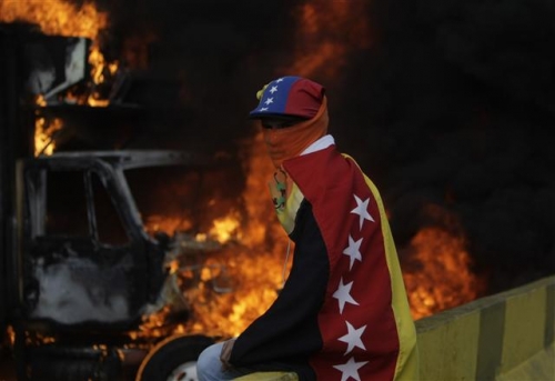 ▲베네수엘라 수도 카라카스에서 27일(현지시간) 한 시위 참가자가 베네수엘라 국기를 몸에 휘감고 화염에 휩싸인 트럭 앞에 앉아 있다. 극심한 경제난을 겪는 베네수엘라에서는 거의 매일 시위가 열리고 있다. 사진=AP뉴시스 
