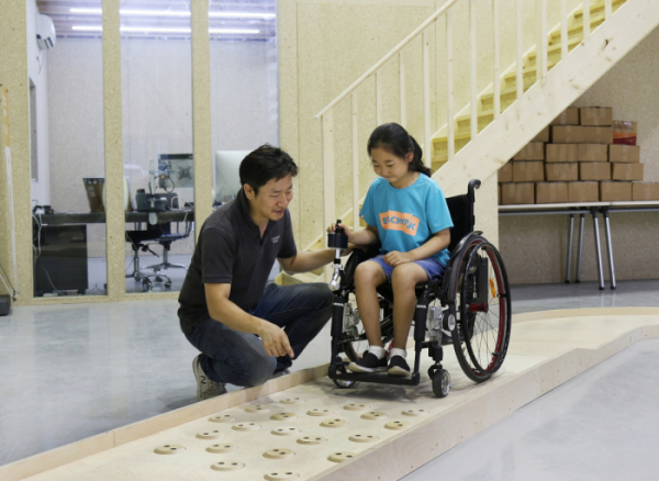 ▲심재신 토도웍스 대표가 토도웍스 사옥에 위치한 휠체어 전용 교육장에서 장애 아동에게 수동휠체어용 전동 키트를 활용한 이동 교육을 진행하고 있다.(사진제공=SK행복나눔재단)