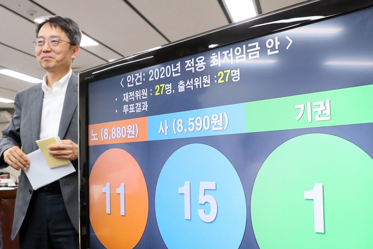 ▲12일 새벽 정부세종청사에서 열린 최저임금위원회 전원회의에서 2020년 최저임금이 2.87% 인상된 8590원으로 결정됐다. 박준식 최임위원장이 결정된 최저임금안 앞을 미소를 지으며 지나고 있다. (뉴시스)