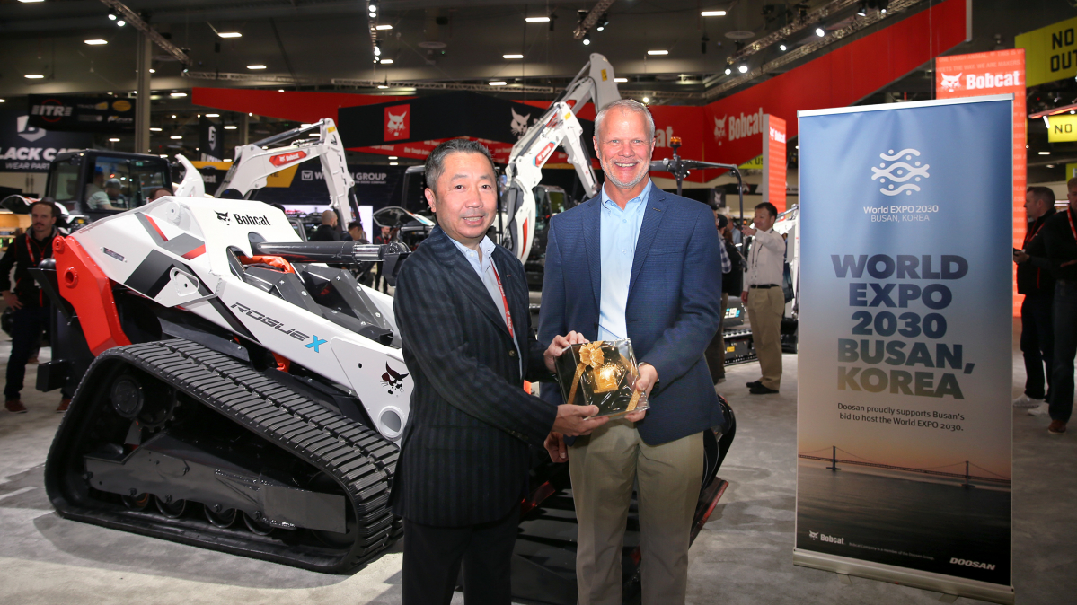 ▲박정원 두산그룹 회장이 미국 라스베이거스에서 열린 ‘콘엑스포 2023’의 두산밥캣 부스에서 무인 전기 콘셉트 로더 ‘로그 X(Rogue X)’ 와 부산엑스포 유치를 기원하는 배너 앞에서 마이크 볼웨버 북미 지역장에게 두산일두(斗山一斗)를 전달하고 있다. (사진제공=두산그룹)