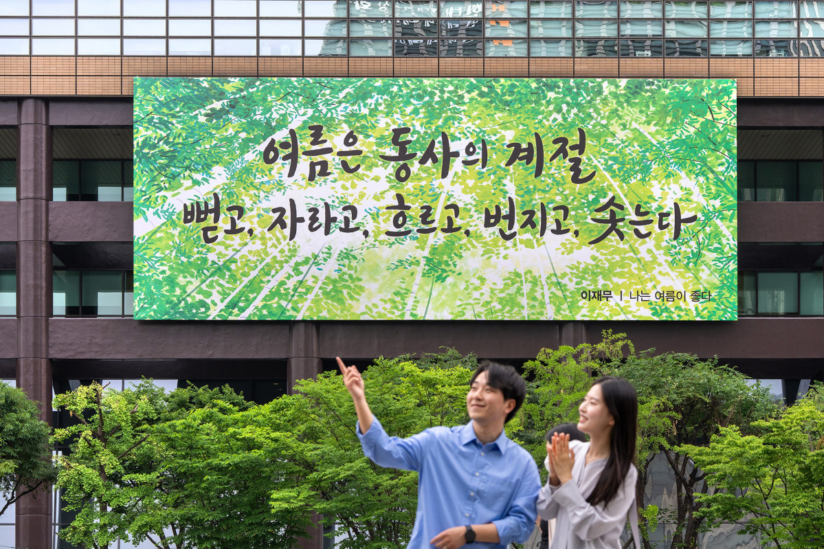 ▲광화문 교보생명빌딩 외벽에 초록색 나뭇잎을 배경으로 이재무 시인의 시 구절이 담긴 대형 포스터가 걸려 있고, 남자와 여자가 포스터 앞에서 남자가 무언가를 가리키고 있다. (사진제공=교보생명)