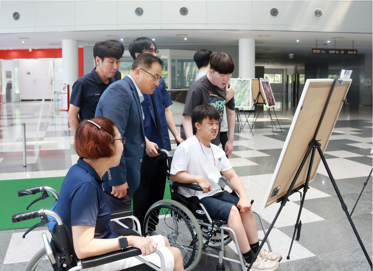 ▲발달장애인 작가 13명의 AI 창작 작품 전시회 '도화지 위의 꿈'에서 휠체어를 이용하는 2명의 작가와 참석자들이 판교글로벌R&D센터 로비에 전시된 작품을 관람하고 있다. (경기도경제과학진흥원)