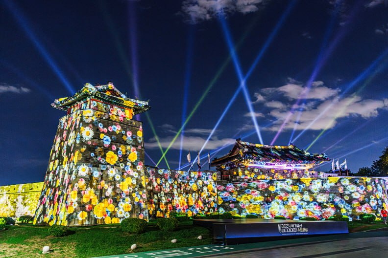 ▲수원화성 성곽에 다양한 꽃들로 구성된 화려한 미디어아트가 투사되고 밤하늘에 다채로운 레이저 조명이 뻗어나가며 역사적인 성곽이 환상적으로 연출되고 있다. (수원문화재단)