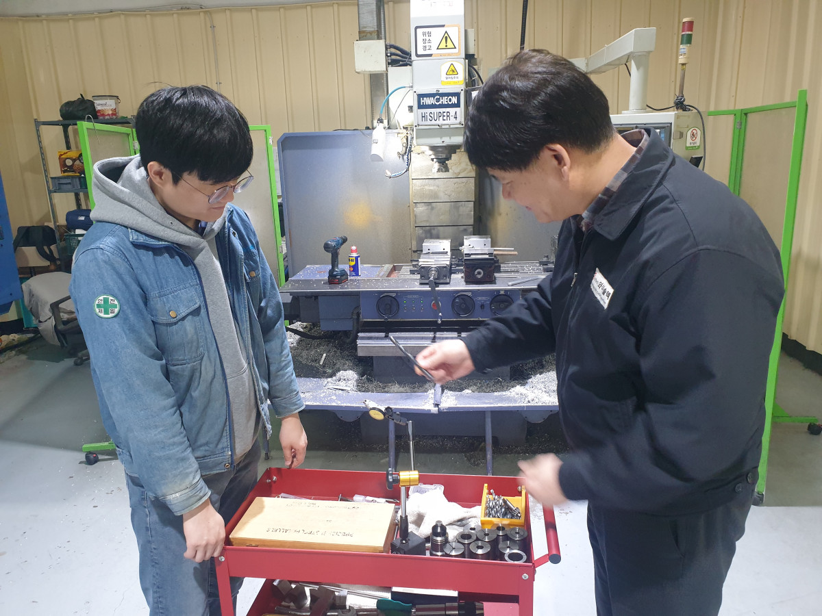 ▲쎄네스테크놀로지㈜ 작업자가 이천시 공장 내에서 HWA-CHEON HI-SUPER-4 기계 앞에서 동료에게 도구 사용법을 설명하고 있다. (경기테크노파크)