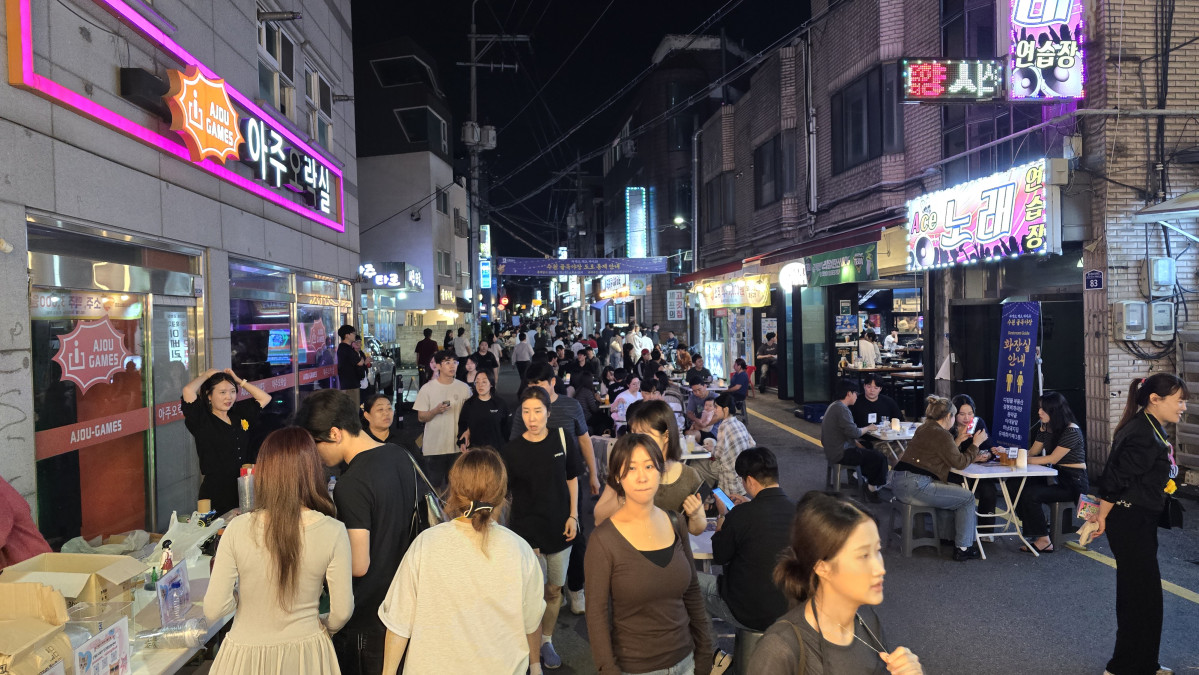 ▲수원 아주대삼거리 인근 아주로4번길의 밤 거리에서 시민들이 상점과 음식점 앞에 모여 축제 분위기 속에 음식을 먹고 이야기를 나누고 있다. (수원도시재단)