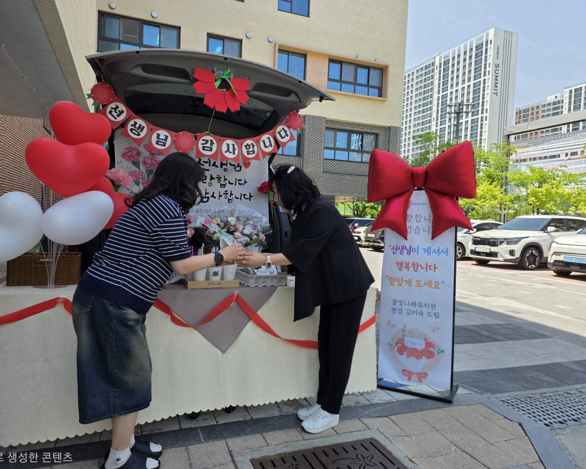 ▲김미숙 물빛나래유치원 원장(오른쪽)이 15일 스승의 날을 맞아 출근하는 교사에게 카네이션 꽃다발을 건네며 감사 인사를 나누고 있다. 뒤편에는 '선생님 감사합니다 사랑합니다'라는 배너와 하트 풍선, 대형 리본으로 장식된 카 트렁크 이벤트 부스가 마련돼 있다. (물빛나래유치원)