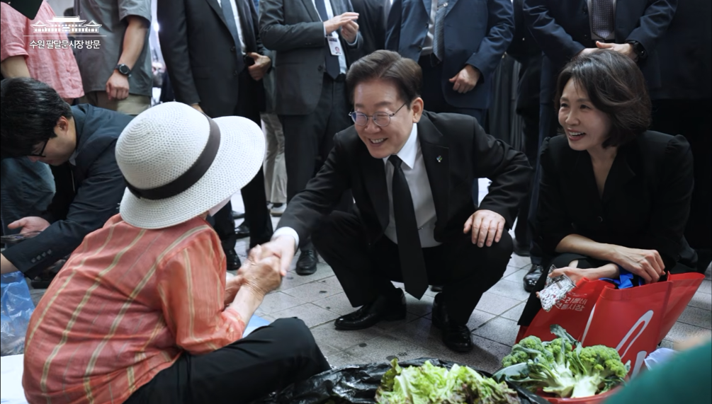 ▲이재명 대통령(가운데) 내외가 25일 수원 팔달문시장에서 야채를 팔고 있는 86세 노인의 손을 잡고 대화하고 있다. 이 대통령은 이 자리에서 노점 단속이 완화됐다는 노인의 말을 듣고 "우리 이재준 시장이 잘하나 보지"라며 이재준 수원특례시장을 호평했다. (이재명TV)